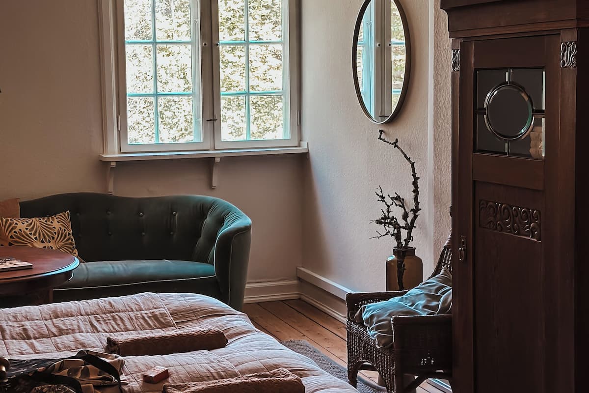 Una habitación con sofá verde, una cama con colcha, un armario de madera y una ventana con vista a los árboles.