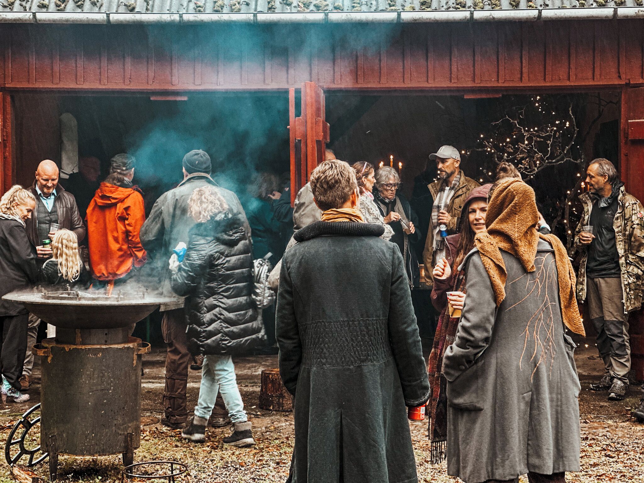 A group of people gathered around a central fire pit in an outdoor setting with a rustic building in the background.
