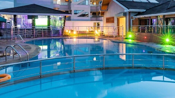 Ein Hallenbad mit klarer Wasserfläche, umgeben von einem Poolbereich mit Geländer und einigen Liegestühlen.