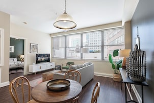 A modern living room with a dining table, chairs, a sofa, a television, and a large window with blinds.