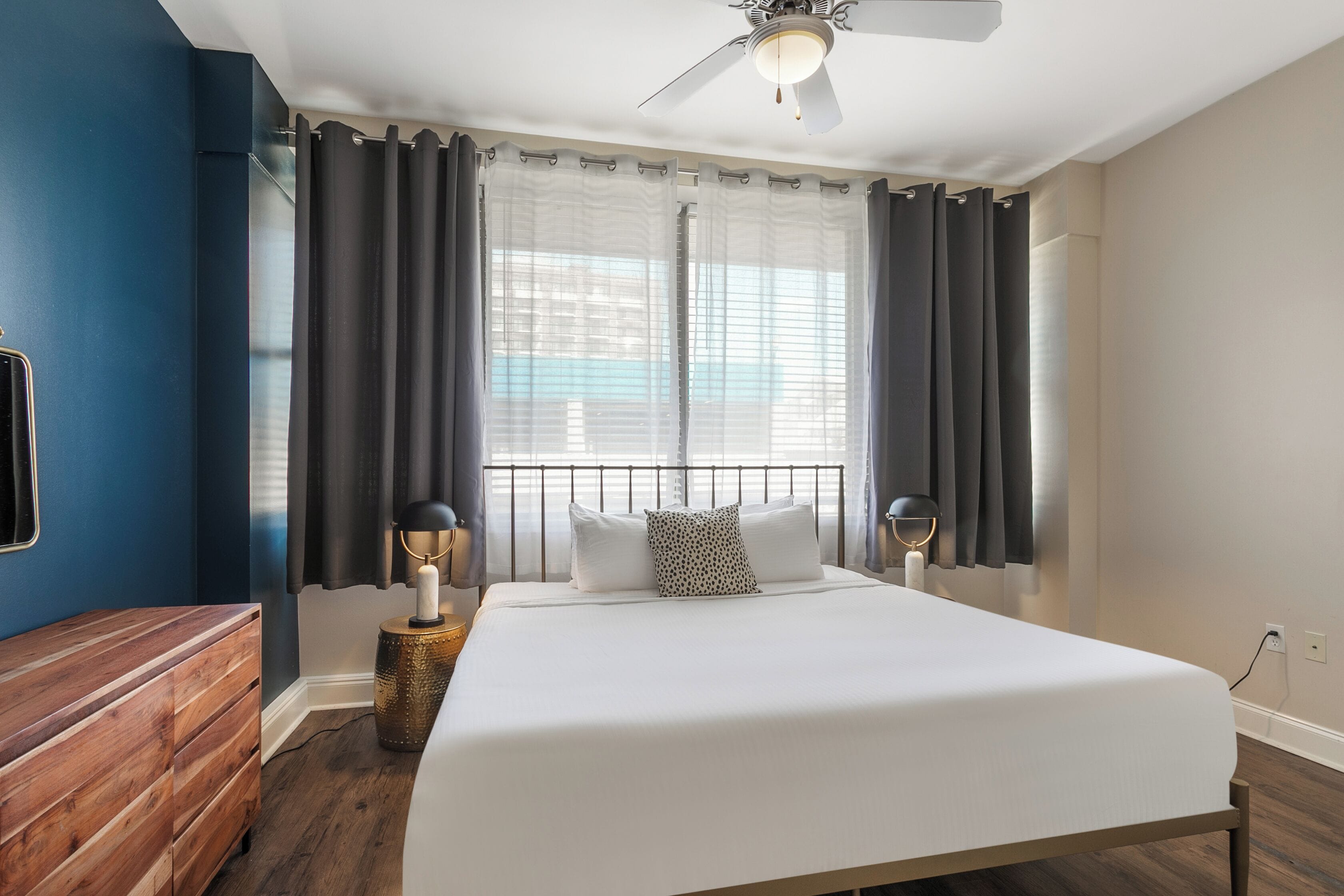 Un dormitorio con una cama grande, cortinas oscuras, ventilador de techo y cómoda de madera.