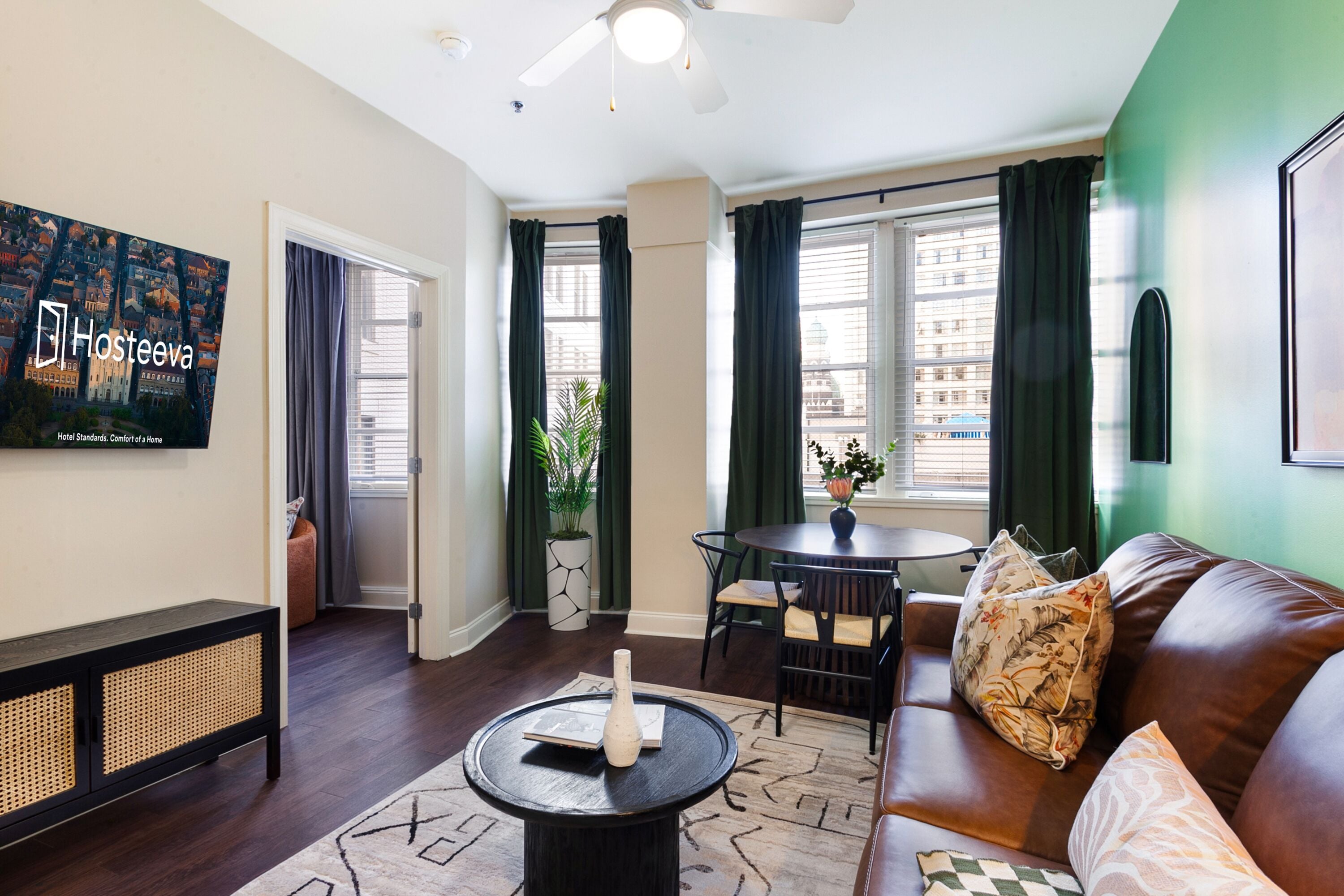 Une chambre d’hôtel moderne équipée d’un canapé, d’une table basse ronde, d’une table à manger avec des chaises, d’une grande fenêtre avec des rideaux, d’un ventilateur de plafond et d’un téléviseur fixé au mur qui affiche le nom de l’hôtel.