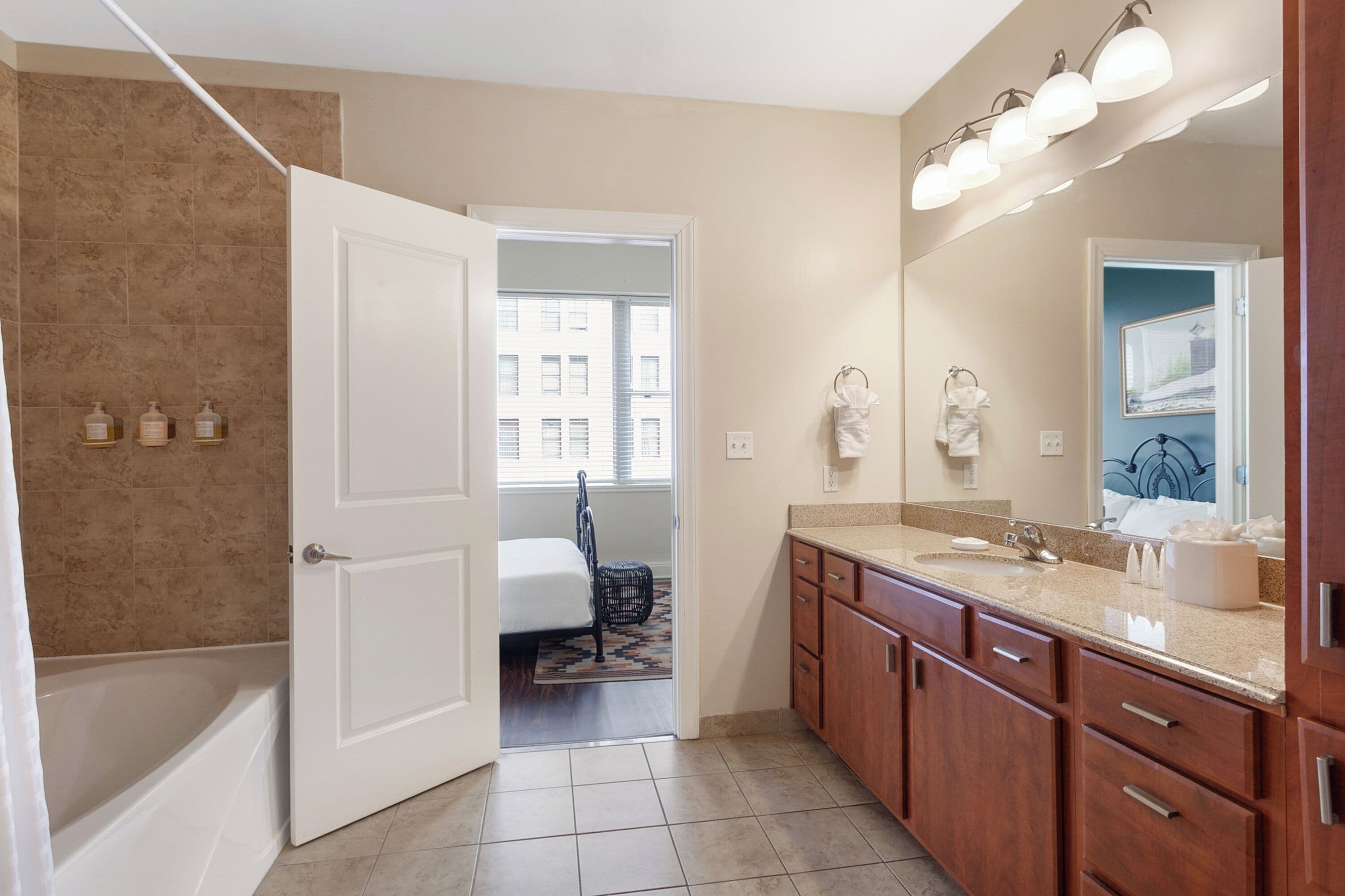 A bathroom with a bathtub, a sink, and a mirror. There is a door leading to a bedroom.