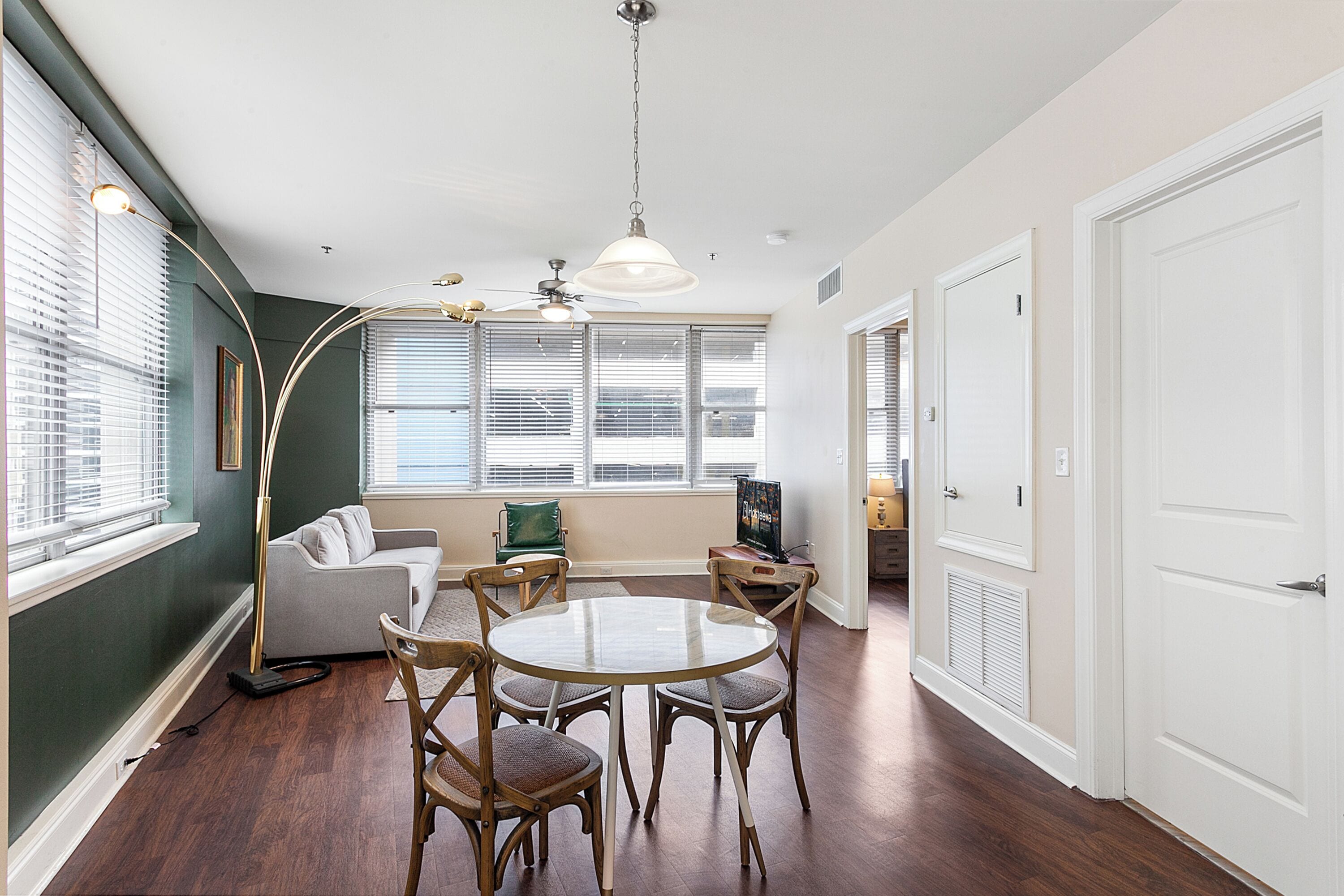 A dining area with a round table and chairs, a ceiling fan, and a large window with blinds.