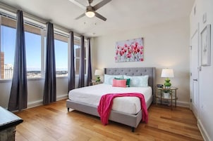 A bedroom with a bed, a ceiling fan, a window with curtains, a painting, and a nightstand with a lamp.
