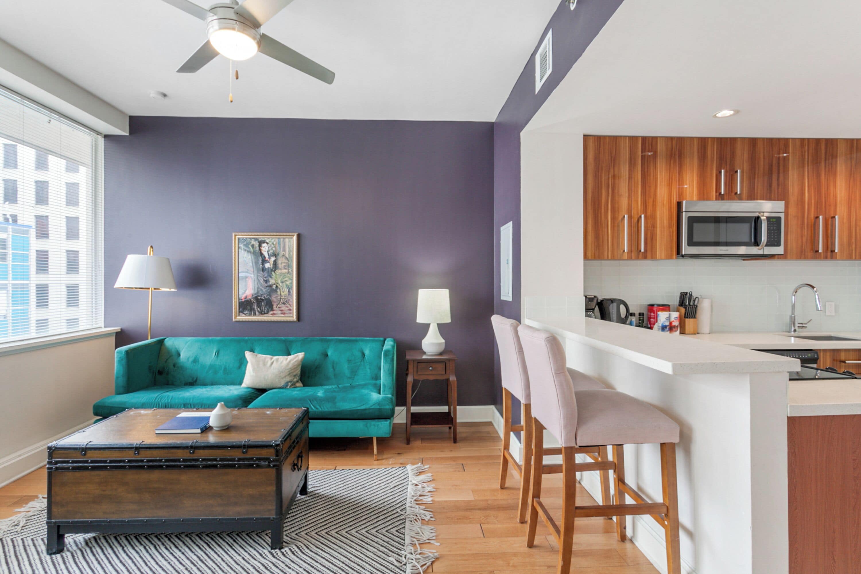 A modern living room with a teal sofa, a wooden coffee table, and a kitchen area with wooden cabinets and a microwave.
