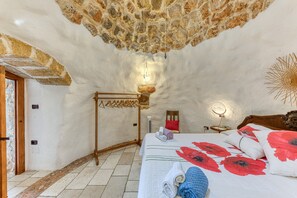 Une pièce avec un plafond en pierre, un lit recouvert d’une couette à motifs floraux rouges, une chaise en bois et un luminaire fixé au mur.