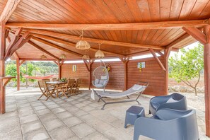 Une terrasse couverte avec des poutres en bois, un hamac, une table avec des chaises, un fauteuil suspendu et un fauteuil de relaxation.