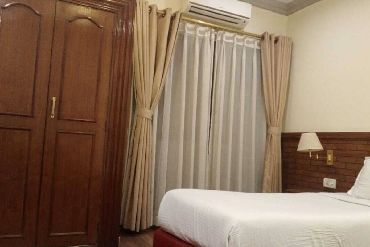 A hotel room with a bed, a wardrobe, curtains, an air conditioning unit, and a wall-mounted lamp.