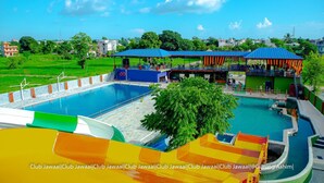 A swimming pool with a slide, covered seating areas, and a view of the surrounding greenery and buildings. - Club Jawai (Butwal)