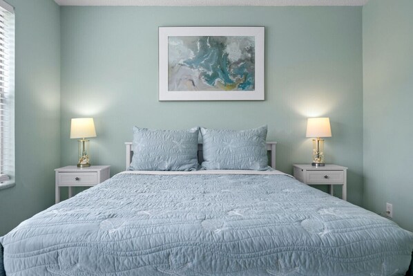 Un dormitorio con una cama, dos mesitas de noche, dos lámparas y un cuadro abstracto enmarcado en la pared.