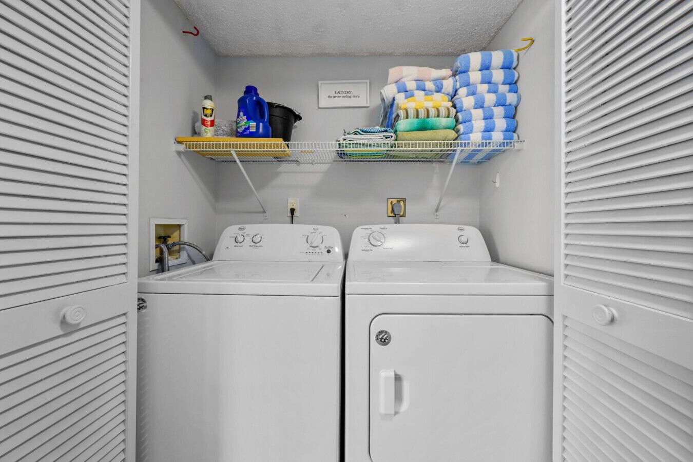 A laundry room with two white front-loading washers and dryers, shelves with cleaning supplies, and a sign above the dryers.