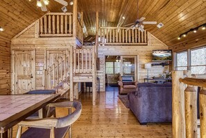 A log cabin interior with a dining area, a living room, and a staircase leading to a loft.