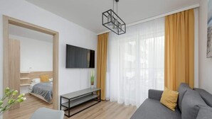 Un salón moderno con un sofá gris, un televisor en la pared, suelo de madera y un amplio ventanal con cortinas amarillas.