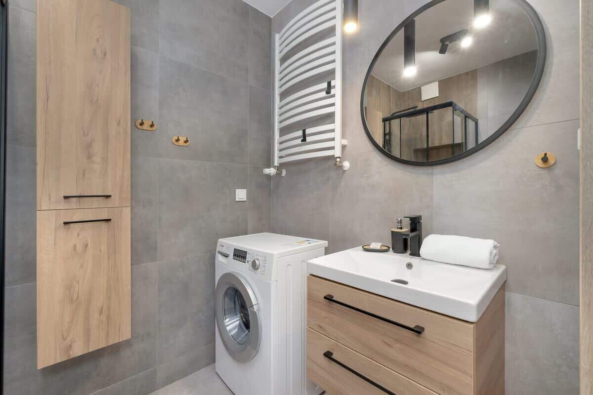 A modern bathroom with a built-in washing machine, a wooden vanity with drawers, a round mirror, and a towel rack.