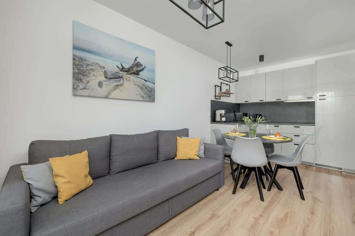 Een moderne woonkamer met een grijze bank, twee gele kussens, een eethoek met een tafel en stoelen, en een keuken op de achtergrond.