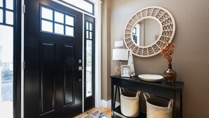 A black front door with a glass panel, a round mirror on a console table, a vase with dried flowers, and two wicker baskets. - Ocean Views! Steps To Beach! 5bd Luxury Home! (Kill Devil Hills)
