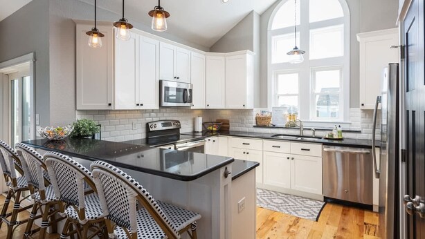 A modern kitchen with white cabinets, a large island, stainless steel appliances, and wicker chairs. - Ocean Views! Steps To Beach! 5bd Luxury Home! (Kill Devil Hills)