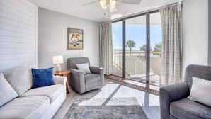 Ein modernes Wohnzimmer mit einem grauen Sofa, einem grauen Sessel und einem blauem Kissen. An der Wand hängt ein Gemälde, ein kleiner Tisch mit einer Lampe steht daneben, und ein großes Fenster ist mit Vorhängen versehen.