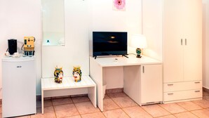 Une pièce de taille modeste, comprenant un bureau blanc, un téléviseur, un petit banc, une armoire et un réfrigérateur.