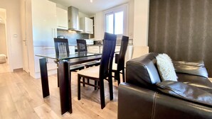 Un salon moderne avec une table à manger en verre, un canapé en cuir noir et un coin cuisine à l’arrière-plan.