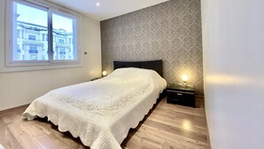 Une chambre avec une grande fenêtre, un lit recouvert d’une courtepointe blanche, une table de chevet et un mur orné d’un motif.
