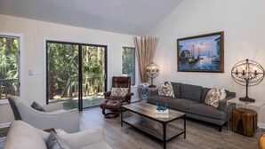 A spacious living room with a sofa, armchair, and coffee table, large windows with a view of greenery, and a painting of a harbor scene. - 723 Marlin Drive (Fripp Island)
