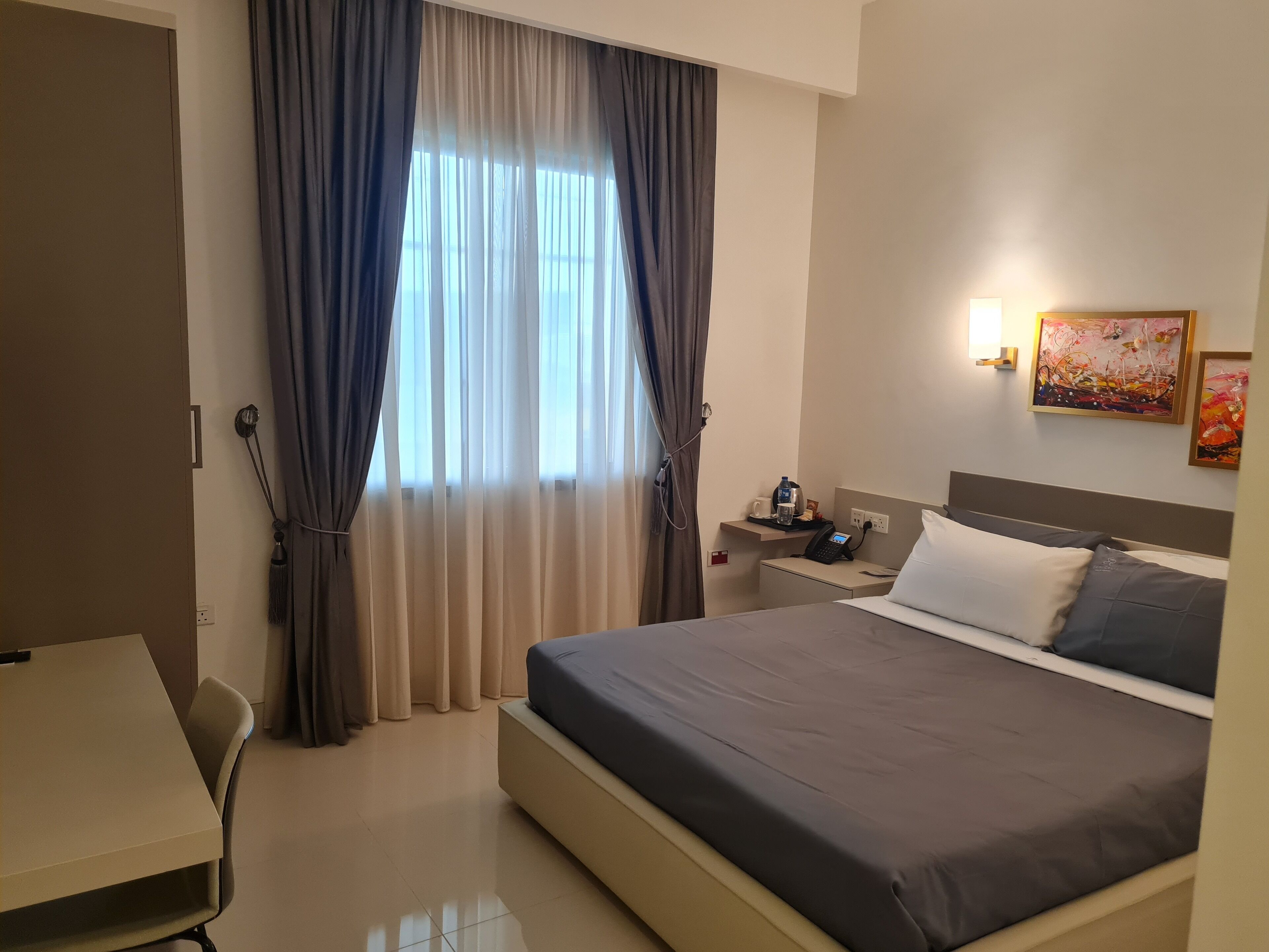 Una habitación de hotel con cama, escritorio, silla, mesita de noche y ventana con cortinas.