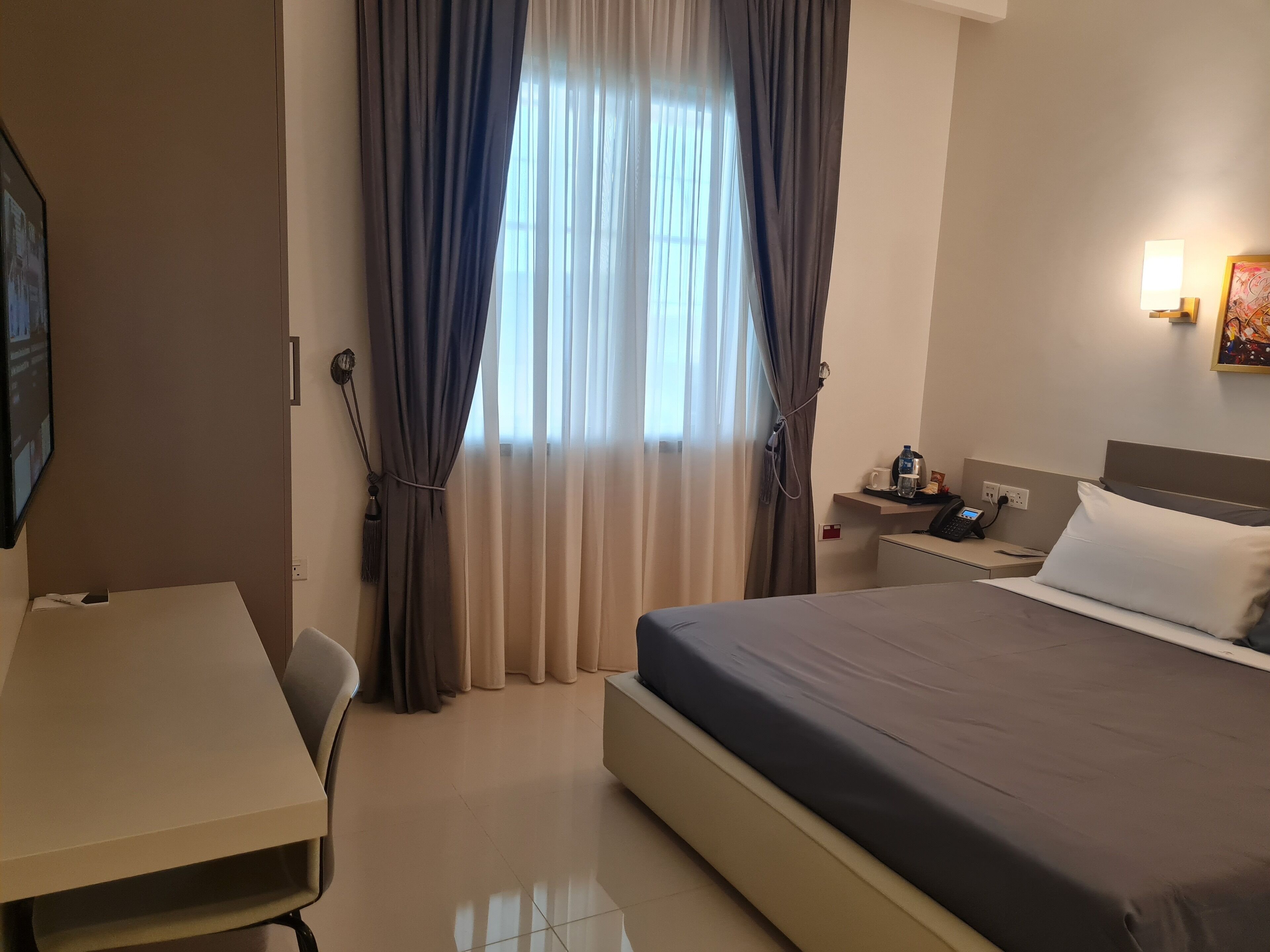 A hotel room with a bed, a desk, a chair, a nightstand with a phone and a lamp, and a window with curtains.
