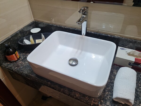 Un lavamanos rectangular blanco con un grifo cromado, un jabonera, una botella de jabón líquido, una caja de pañuelos y una toalla blanca sobre una encimera de granito.