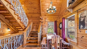 Interior de una cabaña de madera con escalera, mesa de comedor, sillas y una araña.