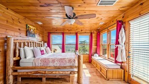 Un dormitorio de cabaña de madera con cama, ventilador de techo, ventanas grandes y vistas al exterior.
