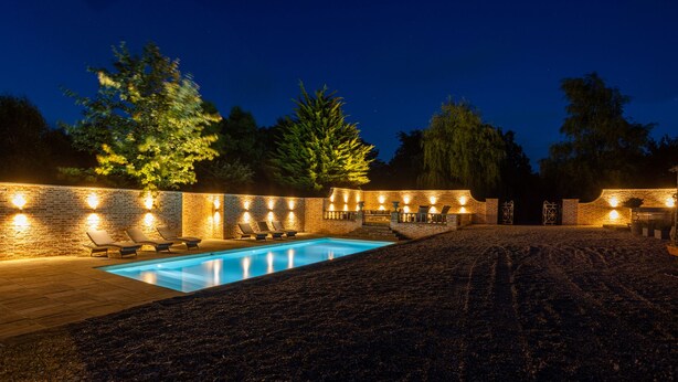 Un espace piscine aménagé pour la nuit, avec un mur en briques, des chaises longues et une piscine.