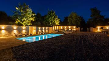 Un espace piscine aménagé pour la nuit, avec un mur en briques, des chaises longues et une piscine.