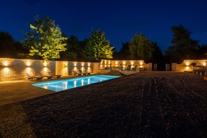 Zona de piscina nocturna con pared de ladrillo, sillones de descanso y una piscina.