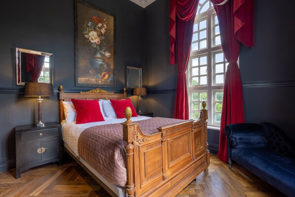 Un dormitorio con cama de madera con dosel, almohadas rojas y blancas, una mesita de noche con lámpara, un aparador y un ventanal con cortinas rojas.