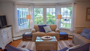 Una sala de estar acogedora con un sofá, sillones, una mesa de centro y un televisor. Amplias ventanas ofrecen vistas al verdor exterior.