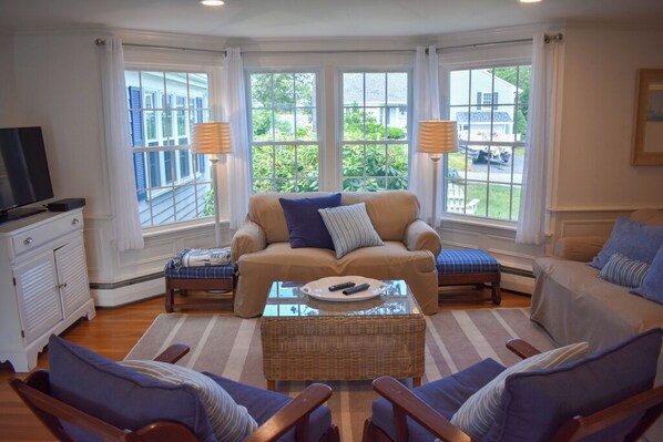 A cozy living room with a sofa, armchairs, a coffee table, and a TV. Large windows offer a view of greenery outside.