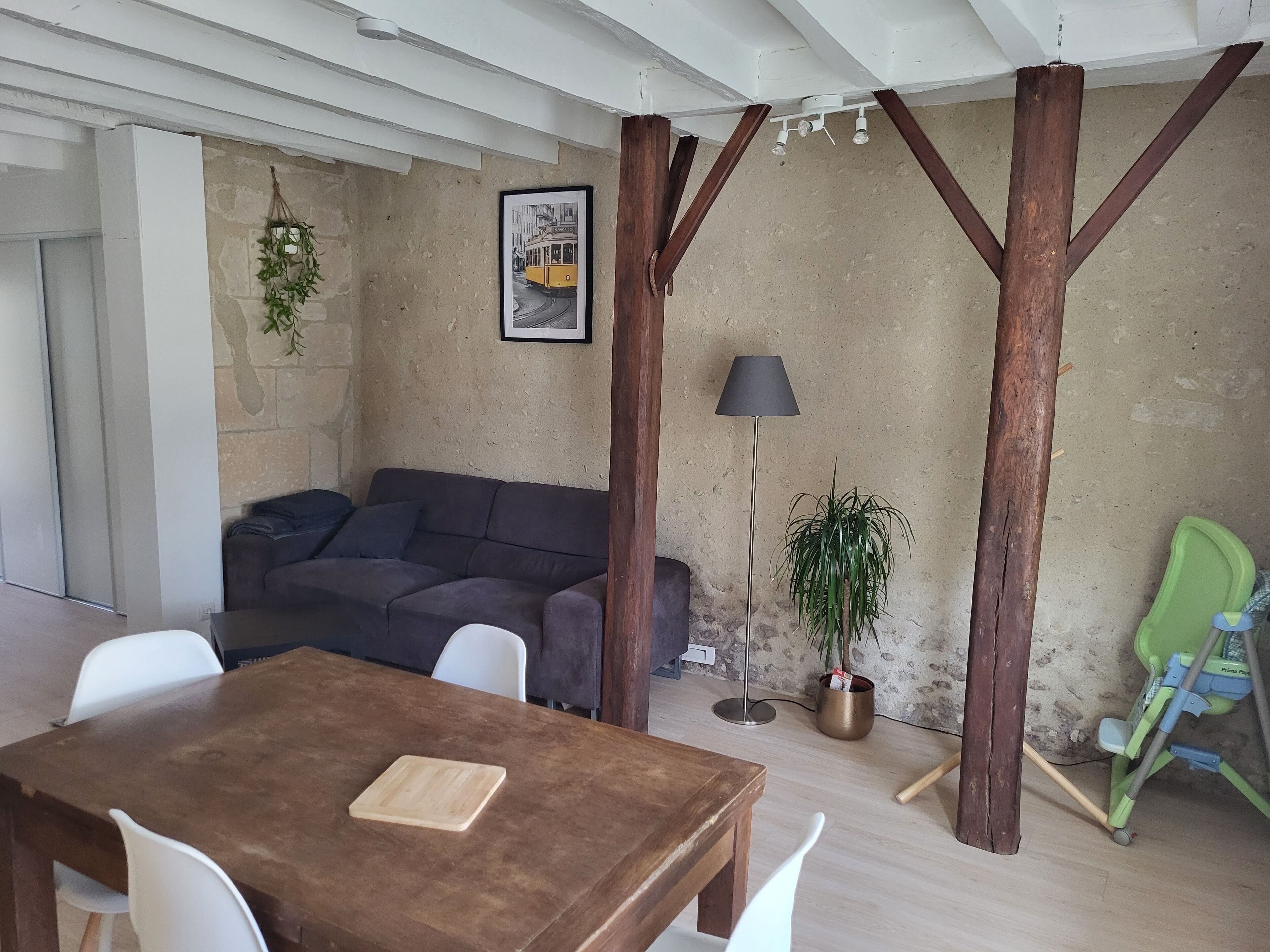 A cozy living area with a wooden table, a sofa, and a dining table. There is a framed picture on the wall and a potted plant.