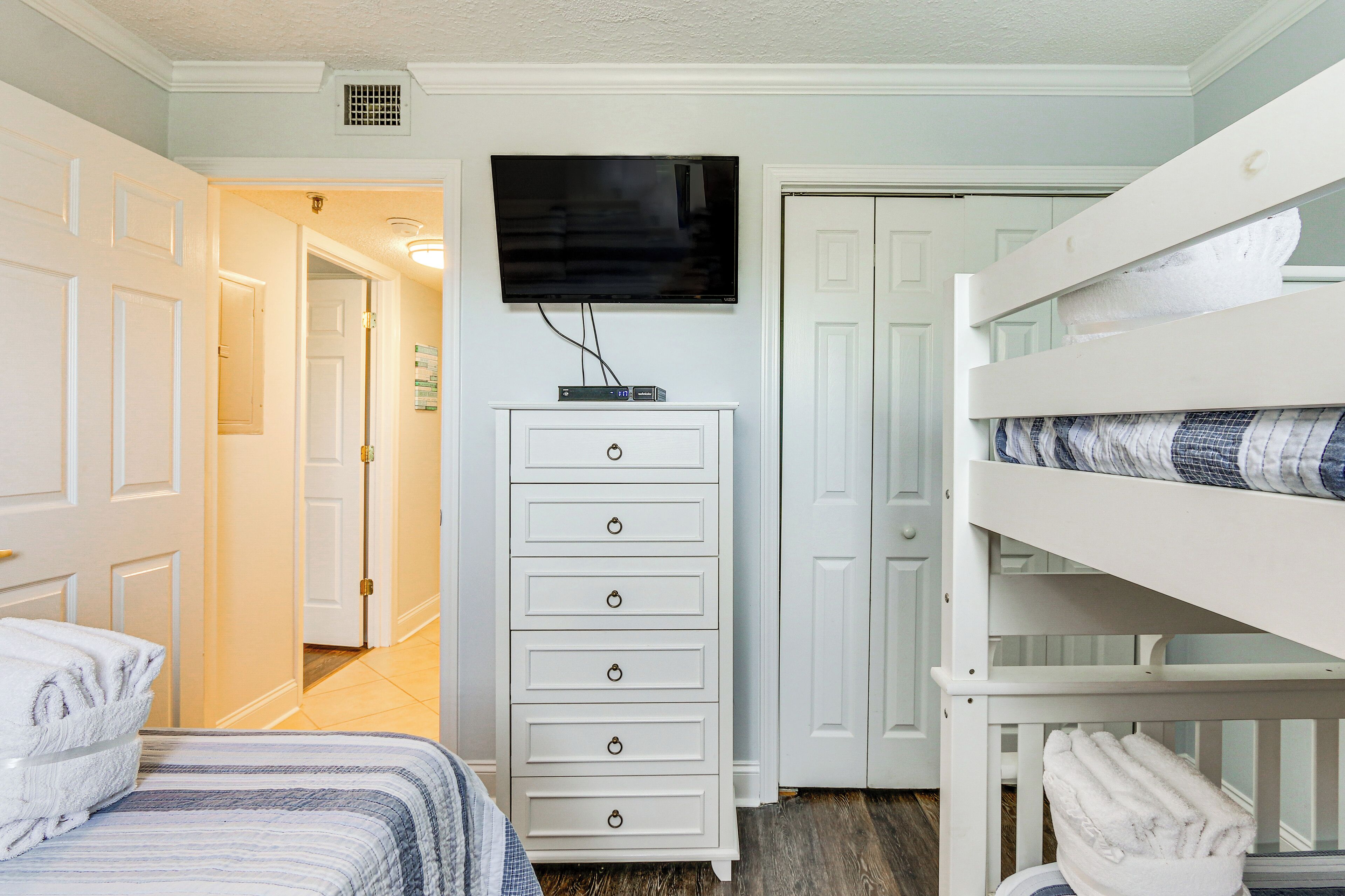 Une chambre avec un lit superposé, une commode blanche, un téléviseur fixé au mur et une porte donnant sur une autre pièce.