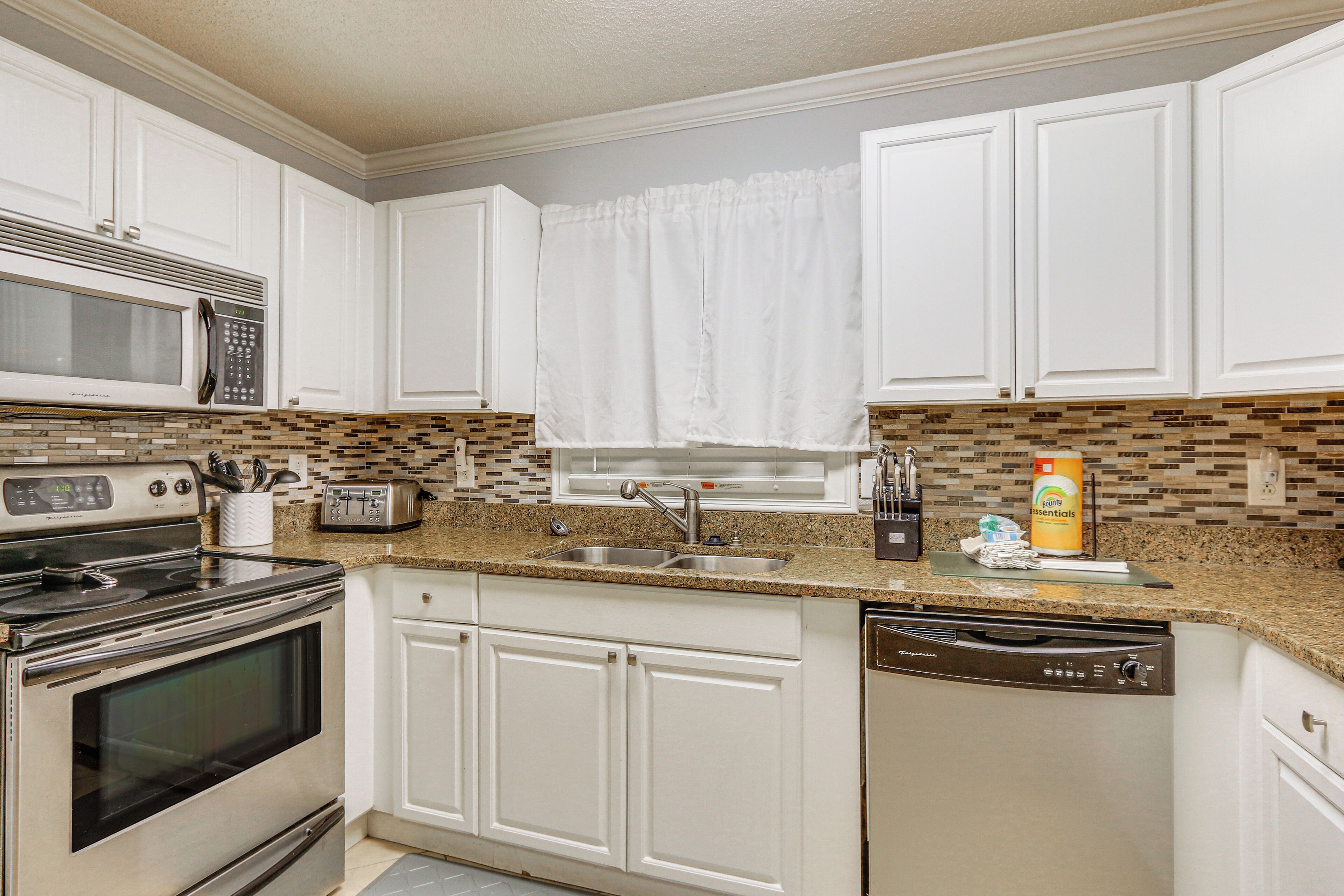A kitchen with white cabinets, a microwave, a stove, a dishwasher, a sink, and a window with a curtain.