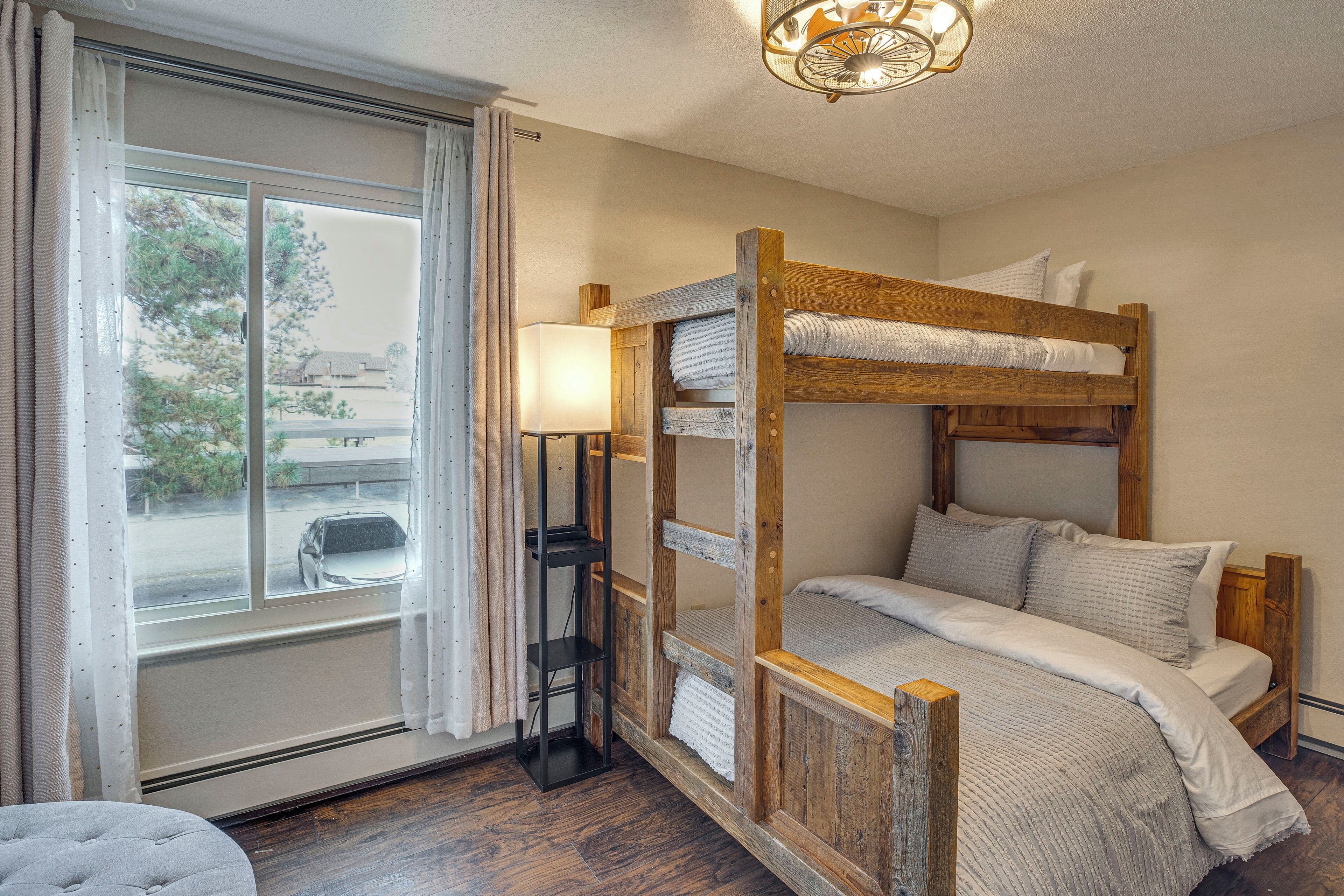 A bunk bed with a wooden frame, a bedside lamp, and a view of the outside through a window with curtains.