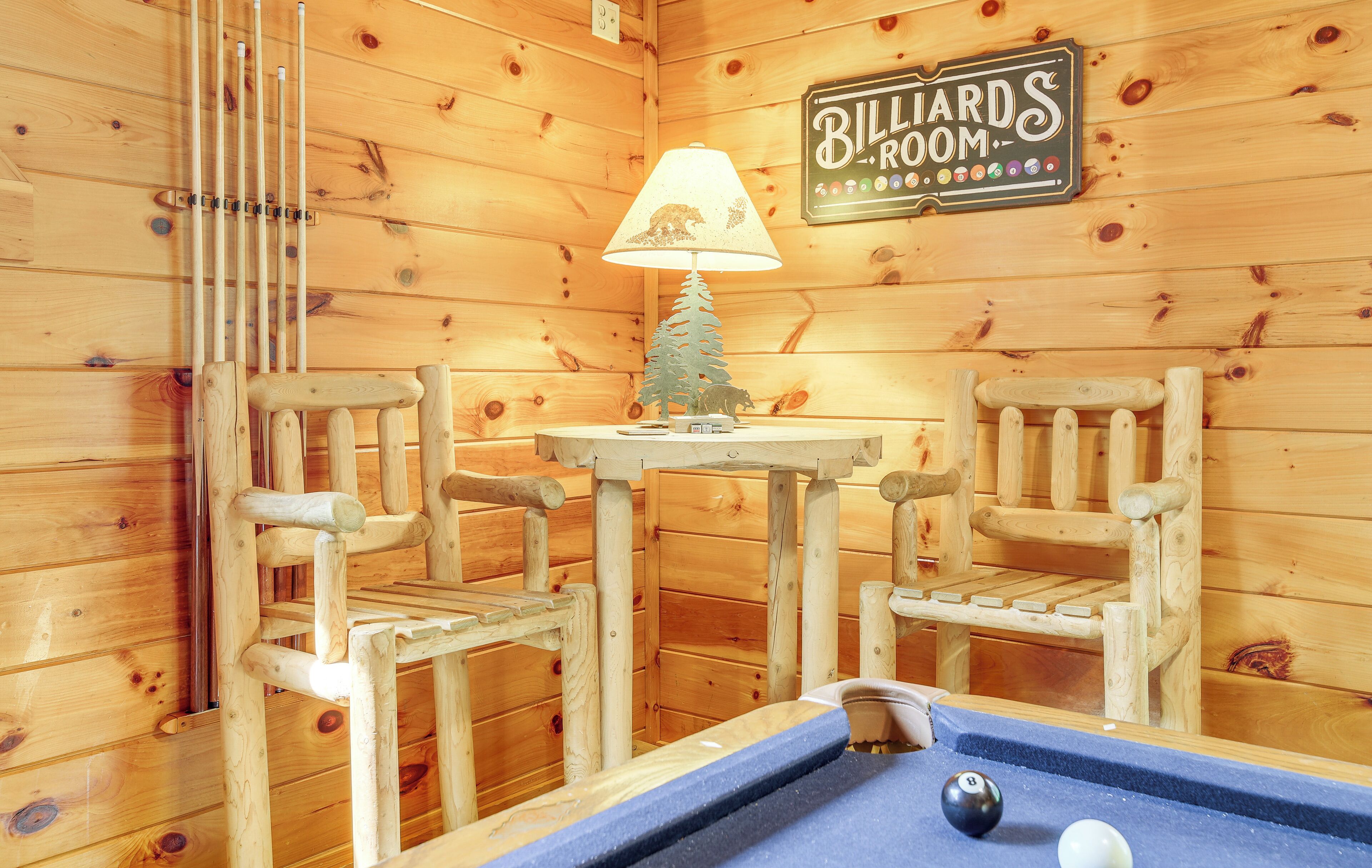 A billiards room with wooden walls, two wooden chairs, a small table with a lamp, and a pool table.