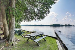 A lakeside picnic area with a wooden bench and picnic table, surrounded by trees and a dock extending into calm water. - Waterfront North Hero Home w/ Screened Porch (North Hero)
