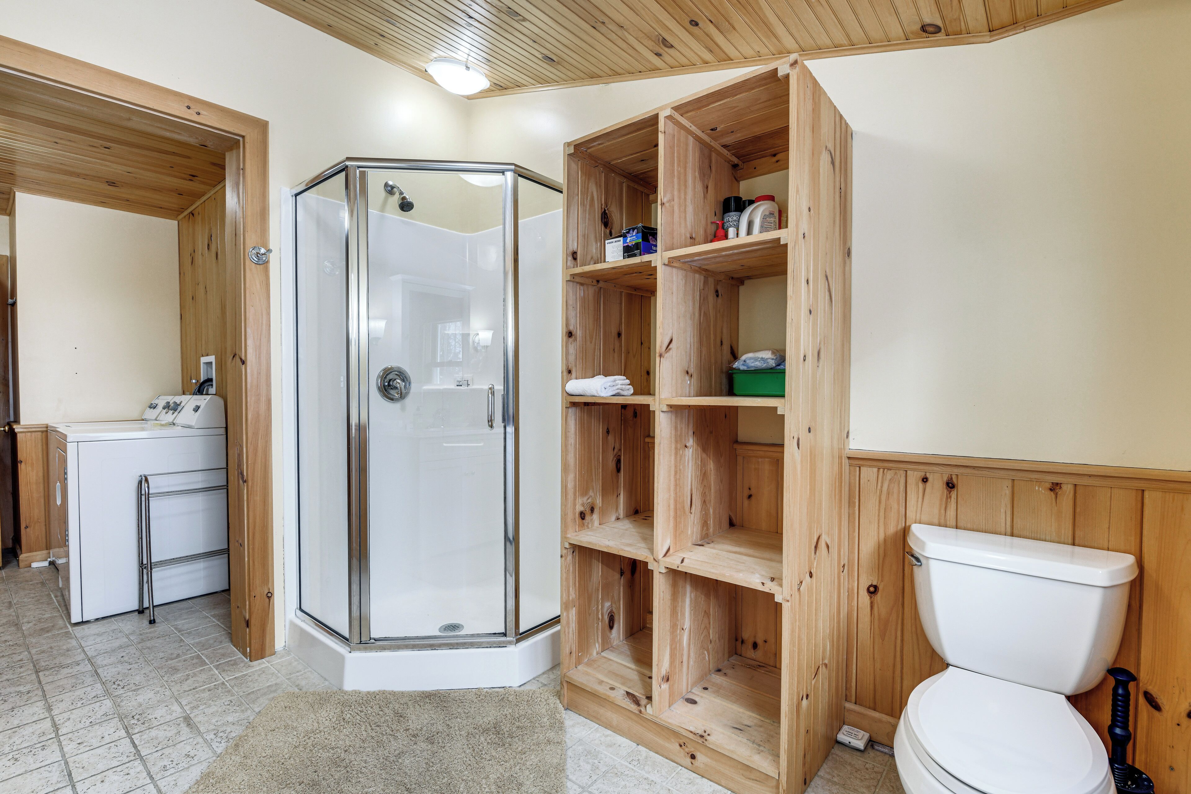 A bathroom with a glass shower enclosure, wooden shelves, a toilet, and a sink.