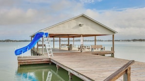 Un muelle de madera se extiende sobre un lago, con una zona de estar cubierta y un tobogán azul.