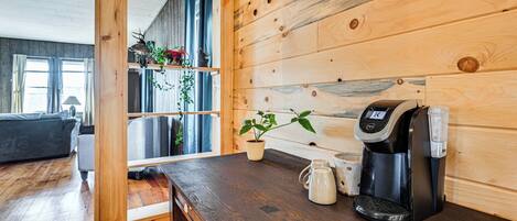 A wooden interior with a coffee maker on a counter, a potted plant, and a view into another room with a couch and a window.