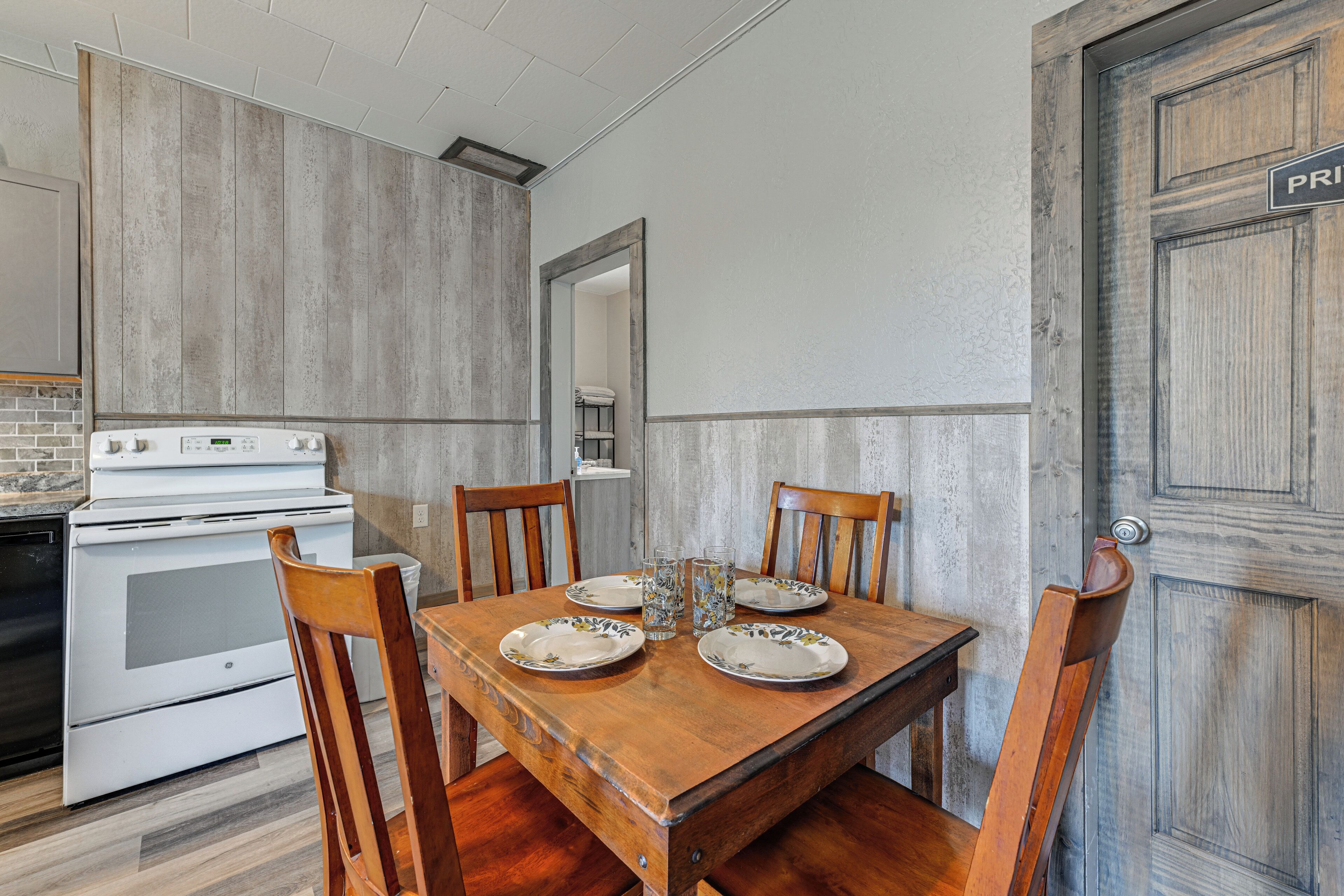 Comedor con una mesa de madera preparada para cuatro personas, rodeada de sillas de madera. Se observa una estufa blanca al fondo.