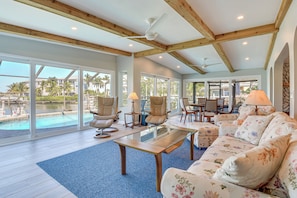 A spacious living area with a glass coffee table, floral sofa, and large windows overlooking a pool.