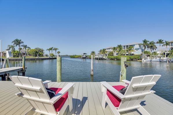 A waterfront deck with two white lounge chairs, a dock, and a marina with boats.